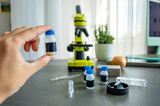 A person holding a small sample vial in front of a yellow microscope, with more scientific supplies on the table. Concept of science education and home experiments for kids