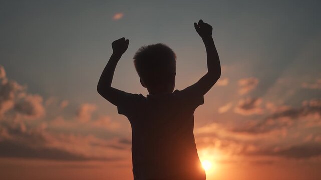 Child silhouette raises arms in sunset light. Boy celebrates freedom high arms in open sky. Arms lifted for victory. Sunset behind boy. Freedom moment for child with arms raised in sunset celebration.