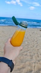 Hand holding an open bottle of orange juice on a sandy beach with the sea in the background. this image conveys refreshment, summer relaxation and outdoor leisure