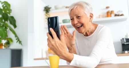 Smiling Senior Woman Waving at Smartphone During Video Call in Bright Kitchen, Digital Communication, Lifestyle. Mobile Device. Internet Connectivity, Tech Engagement. Useful for Editorial.Copy Space