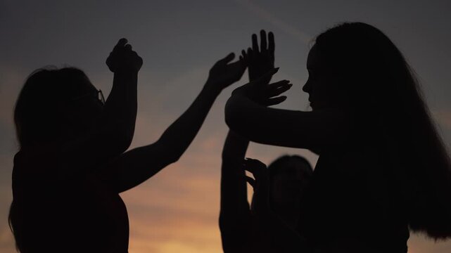 People raising hands in silhouette against sunset sky, group celebrating and dancing at dusk, hands reaching, silhouette profile, party vibe, friendship energy and joyful movement under warm orange