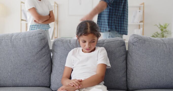 Child watches parents quarrel during family conflict. A sad girl on the sofa sits, head down as parents argue at home. Trauma and stress from argument show. Divorce and conflict harm children.