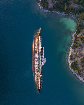 Aerial view of the rusted MS Mediterranean Sky wreck lying in crystal blue coastal waters as the sun sets over the shoreline of Athens, Greece.