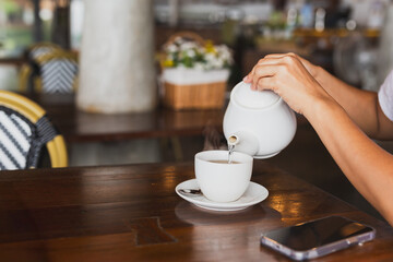 Female hand pouring from white ceramic teapot into a cup.