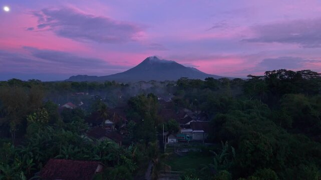 Aerial view of the imposing Mount Merapi, towering above a lush forest under a vibrant purple and pink sky, Klaten Regency, Central Java, Indonesia.