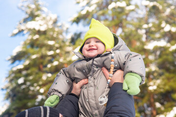 Portrait of a happy baby boy in a snowy forest on a sunny day. Dad lifts the baby up. The child is 10 months old.