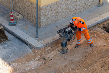 Construction worker in orange safety gear operating a tamping rammer machine to compact gravel in a trench. Road maintenance scene with an excavator bucket and traffic cone nearby.