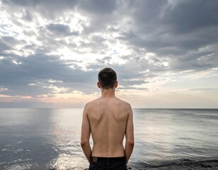 A shirtless man stands on a rocky shore, facing a calm sea under a cloudy, soft-lighted sky