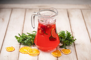 Red citrus lemonade in a glass pitcher with mint, orange slices and fresh herbs on rustic wooden background