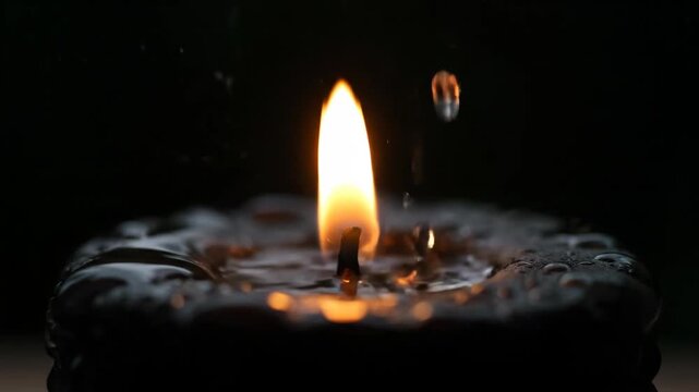 A black candle burns as water drops fall in slow motion. Close-up of a flickering flame splashing in dark wax. Abstract concept of fire and water elements