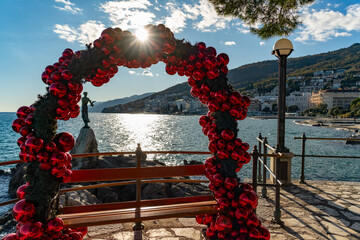 Festive red ornament arch framing a seafront bench with a view of the Opatija coastline and the “Maiden with the Seagull” statue. Bright sunlight and sparkling sea create a warm holiday atmosphere