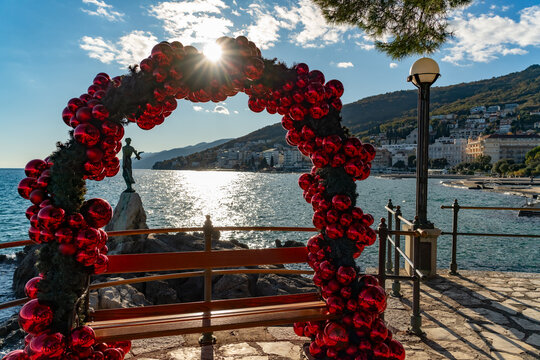 Festive red ornament arch framing a seafront bench with a view of the Opatija coastline and the “Maiden with the Seagull” statue. Bright sunlight and sparkling sea create a warm holiday atmosphere