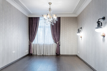 elegant empty room with wooden brown floors, wallpaper and retro lamps on wall, and chandelier with glass pendants on ceiling. room has cozy dim light from lamps. beautiful heavy curtains on window