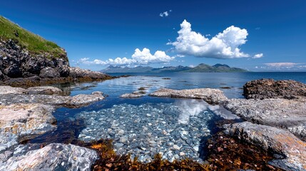 A clear, shallow tidal pool filled with smooth pebbles, set against a rugged, rocky coastline. The calm water reflects the bright blue sky and fluffy white clou