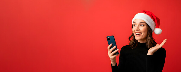 Happy woman in Santa hat looking with smile at mobile phone in Christmas holiday. Communication on smartphone in holiday in studio on red background, banner