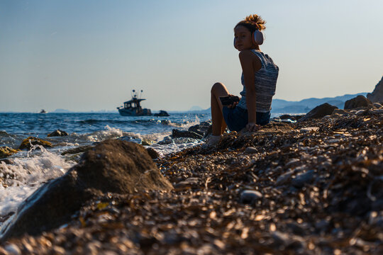 Young girl on beach listening to music. She looks directly at the camera with an anchored speedboat in the background. Summer portrait.