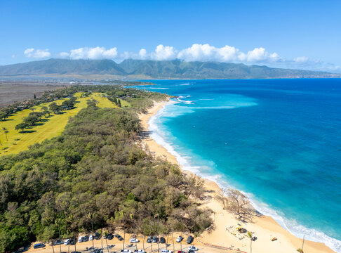 Aerial view of the golden sands meeting the turquoise waters, fringed by lush greenery, under the watchful gaze of distant mountains, Maui, Hawaii, United States.