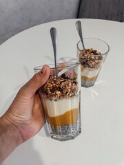 Hand Holding Chia Seed Parfait with Granola and Mango. A person's hand holds a glass of layered healthy dessert or breakfast, featuring chia seed pudding, a bright mango or pumpkin layer, yogurt