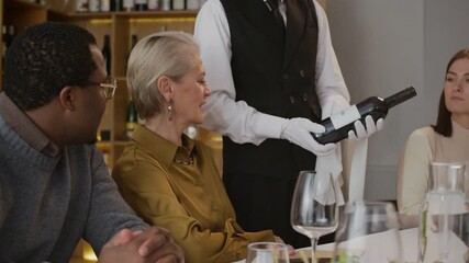 Midsection shot of unrecognizable male sommelier in classic outfit presenting bottle of wine showing its label and sharing his knowledge with multiethnic group of people attending degustation event