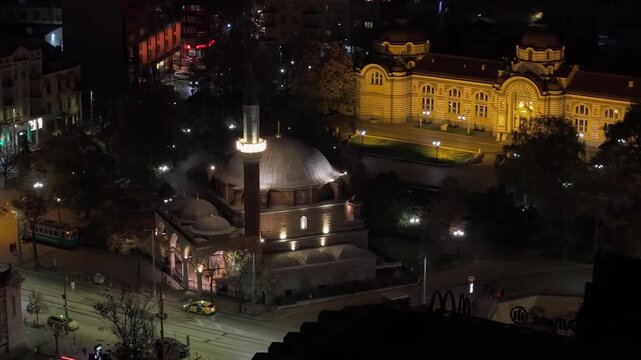 Drone orbit at night around the illuminated Banya Bashi Mosque, revealing the glowing Sofia Regional History Museum in the background.