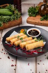 Golden crispy spring rolls served with soy dip on black plate surrounded by herbs and spices