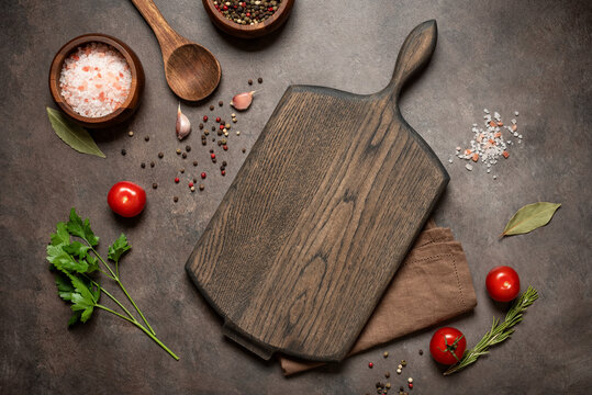 Culinary background with empty wooden cutting board, wooden spoon and spices, dark grunge background, top view, flat lay, copy space.