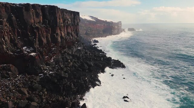 Aerial view of storm ocean waves crashing against snowy coastal cliffs at winter sunrise. The Icelandic plateau reveals striking, martial red mineral cliffs contrasted against the icy landscape