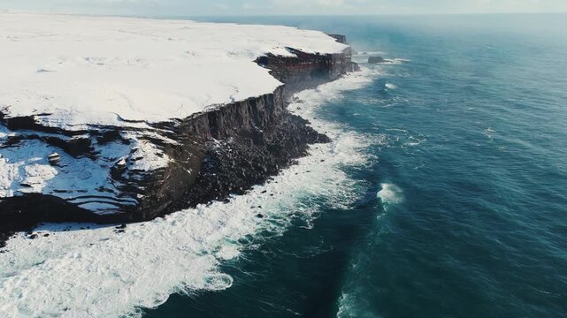 Aerial view of storm ocean waves crashing against snowy coastal cliffs at winter sunrise. The Icelandic plateau reveals striking, martial red mineral cliffs contrasted against the icy landscape