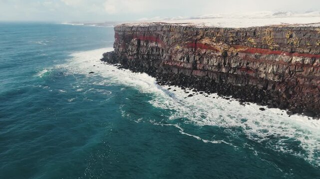 Aerial view of storm ocean waves crashing against snowy coastal cliffs at winter sunrise. The Icelandic plateau reveals striking, martial red mineral cliffs contrasted against the icy landscape