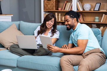 Smiling couple sits on a sofa comparing a tablet and laptop, coordinating tasks for budgeting, study, or a small business project. Real home collaboration, remote work energy, positive teamwork.