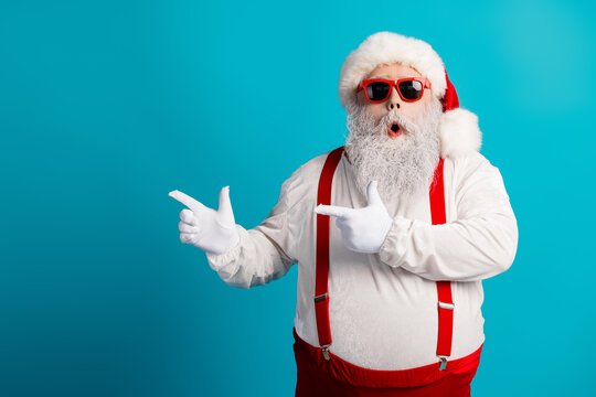 Santa Claus in a thick white beard red suspenders and white gloves wears a hat and cool sunglasses against a bright blue background - Powered by Adobe