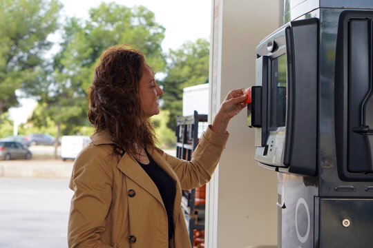 A woman with curly hair and a brown trench coat, smiling as she pays with her card at a gas station
