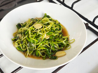 Close-up of sunflower sprouts stir-fried with oyster sauce, garnished with sliced garlic. The vibrant green color contrasts beautifully against the white plate, healthy cooking.