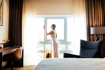 Woman with suitcase standing by hotel window taking picture of city view.