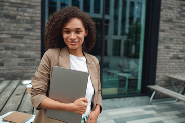 young black woman holding laptop against chest, standing outside modern building, poised expression, campus