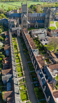 Aerial view of Vicar's Close, a medieval street with honey-colored stone buildings and vibrant green gardens casting long shadows under a clear sky, Wells, England, United Kingdom.