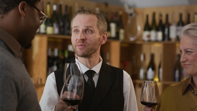 Inspired middle-aged Caucasian male sommelier in elegant outfit teaching art of wine tasting to diverse group of gourmet people swirling glasses and smelling it before taking sip