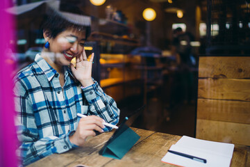 Woman using digital tablet with stylus at a café window, engaged in mobile creative work and personal tech-driven workflow while reflecting city life.