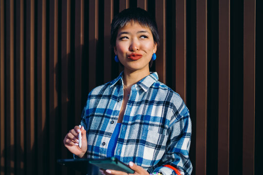 Young Asian woman making a quirky face with lips puckered while holding stylus and tablet, merging playfulness with productivity in personal use of digital technology.