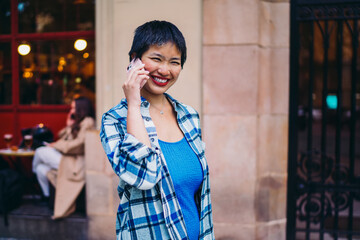 Asian woman on smartphone call outdoors, laughing confidently while holding phone to ear,...