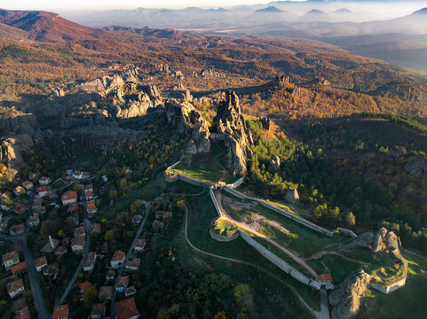 Aerial view of sunlit Belogradchik Fortress nestled amidst rugged rock formations and autumnal forests, a tapestry of ochre and green, Belogradchik, Vidin, Bulgaria.