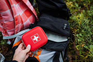 hand holding red first aid kit over backpack on grass, white cross visible, plaid jacket nearby,...