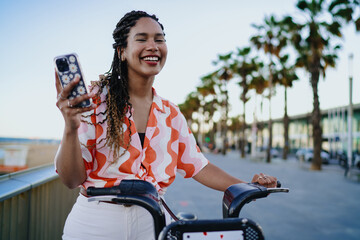 Smiling young woman with braids holding smartphone while resting on red rental bike, symbolizing digital lifestyle, modern mobility and joyful independence in everyday technology use.