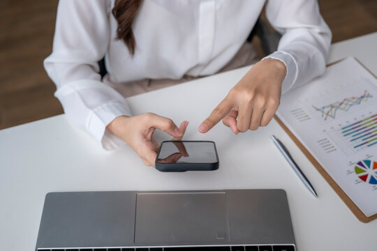 Businesswoman using smartphone and pointing at it with charts and laptop on desk