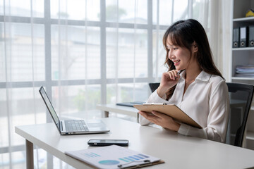 Businesswoman working at office desk using laptop and taking notes on clipboard