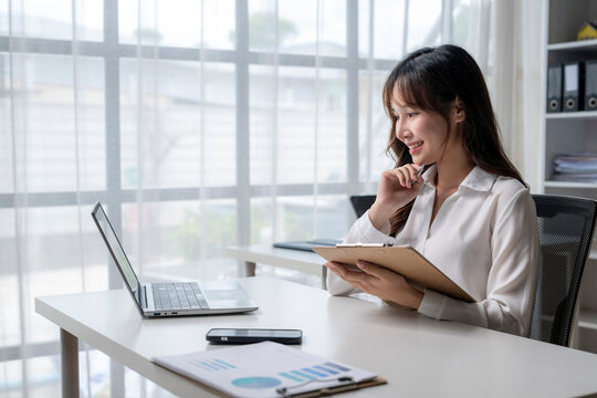 Businesswoman working at office desk using laptop and taking notes on clipboard - Powered by Adobe