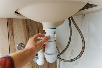 hand reaching under wooden sink basin to tighten trap and check connections, visible braided hoses and cabinet base, handson plumbing maintenance scene for home repair and service