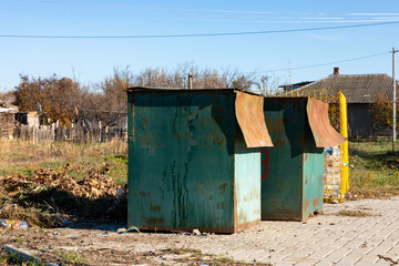 Two old metal garbage bins with rusted lids on a paved area outdoors, village houses and dry grass in the background under clear blue sky.