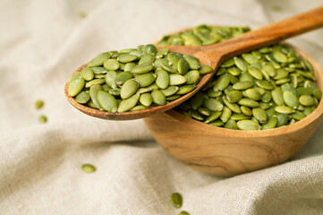 Raw peeled green pumpkin seeds in wooden spoon on natural linen fabric background top view. Food background, Organic healthy tasty pumpkin seeds, Autumn cooking ingredient. Plant-based protein source