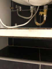 Detailed view of the bottom part of a domestic water heater with visible pipes, fittings, electrical cable, and cabinet shelf showing wear, suitable for concepts of maintenance, repair, and home